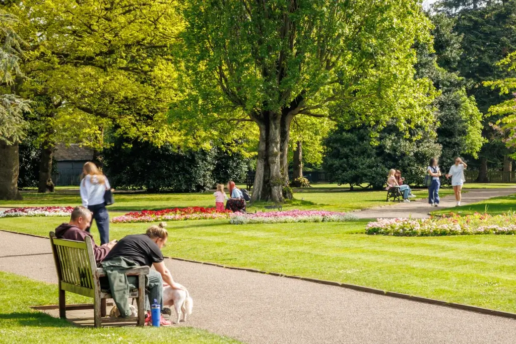 Park scene with people walking, benches, flowerbeds and large leafy trees on a sunny day.