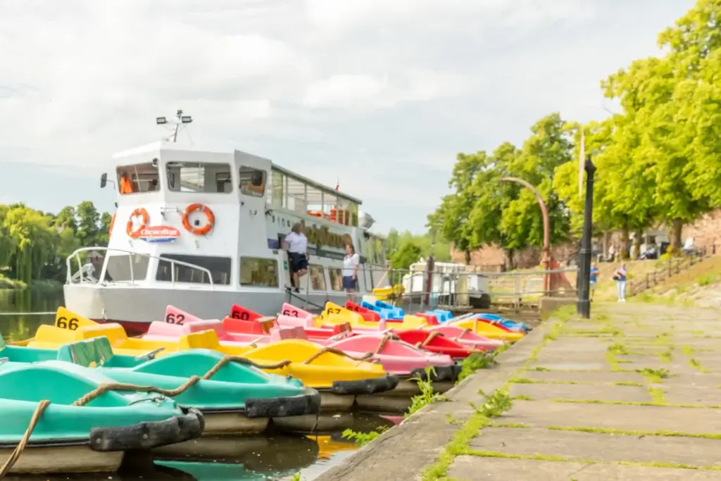 Colourful pedal boats lined up at a quay with a white sightseeing boat docked and leafy trees along the riverbank.
