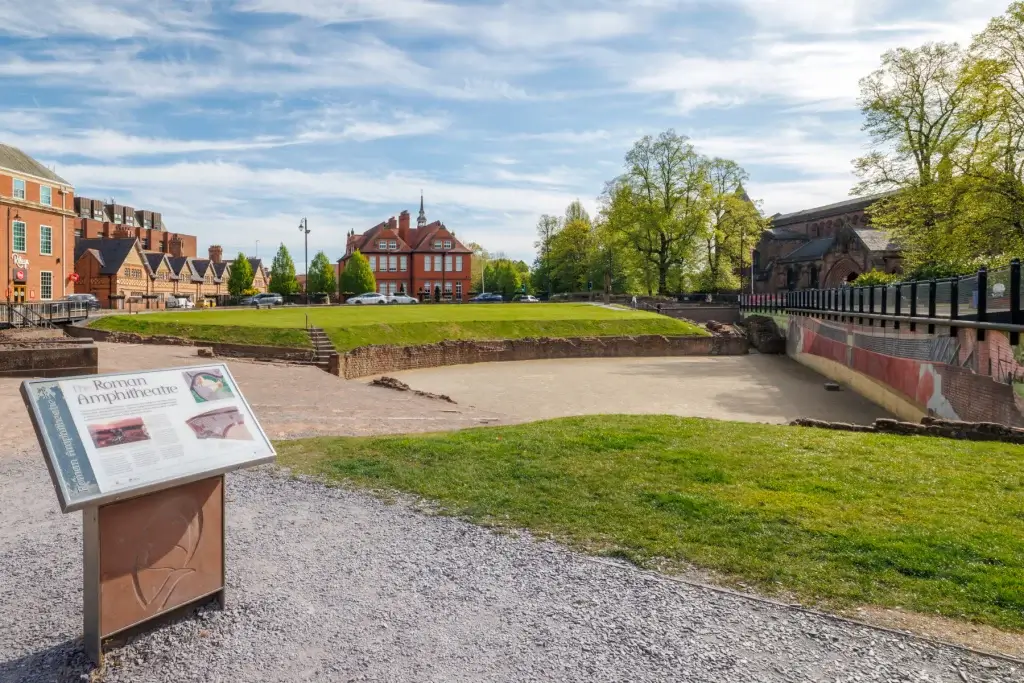 Open grassy archaeological site labeled "The Roman Amphitheatre" with informational sign in foreground and red-brick buildings and trees beyond