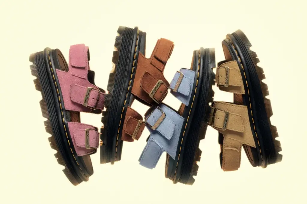 Four suede Dr. Martens sandals in pink, brown, blue and tan shown side-on with chunky black soles and metal buckles.