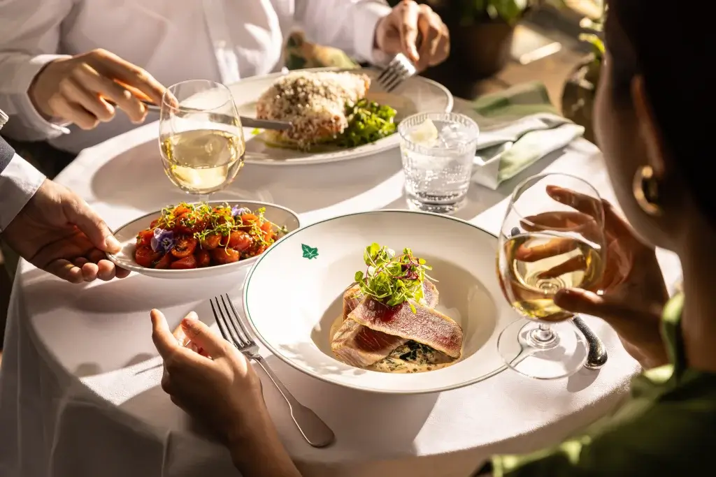 Seared tuna on cream-spinach sauce topped with microgreens on a white plate, with wine glasses, tomato side and diners' hands.