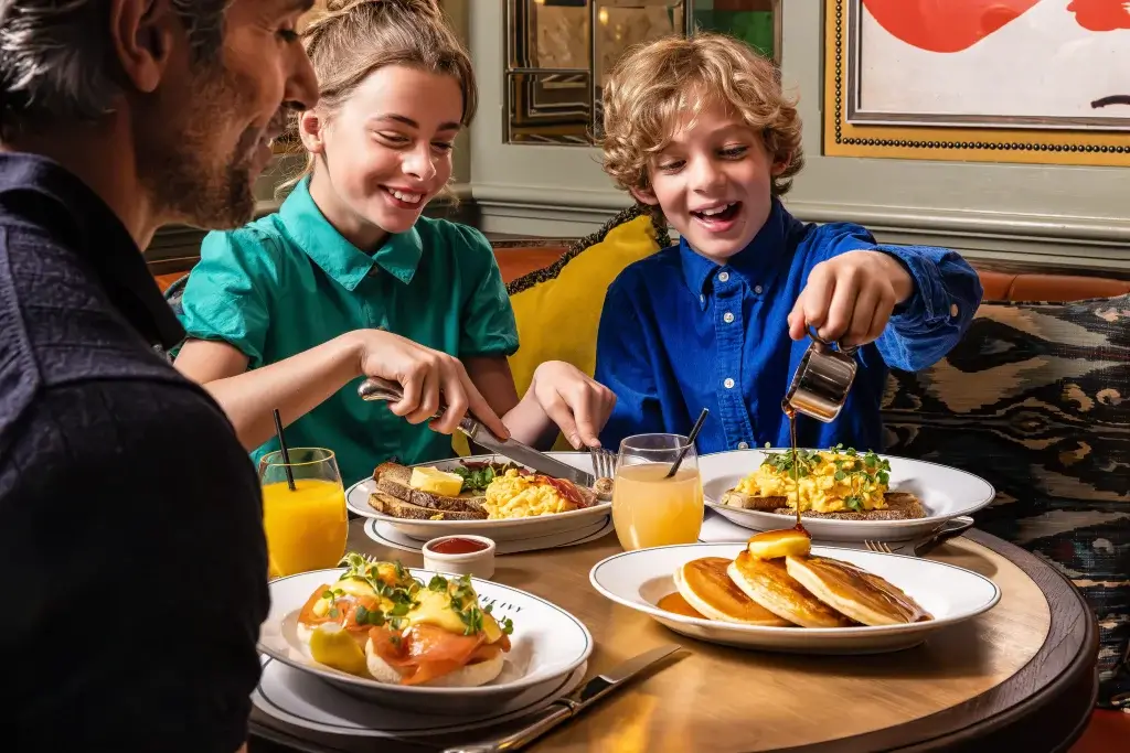 Smiling children at a table pouring syrup and cutting food while enjoying a breakfast with pancakes, scrambled eggs and orange juice