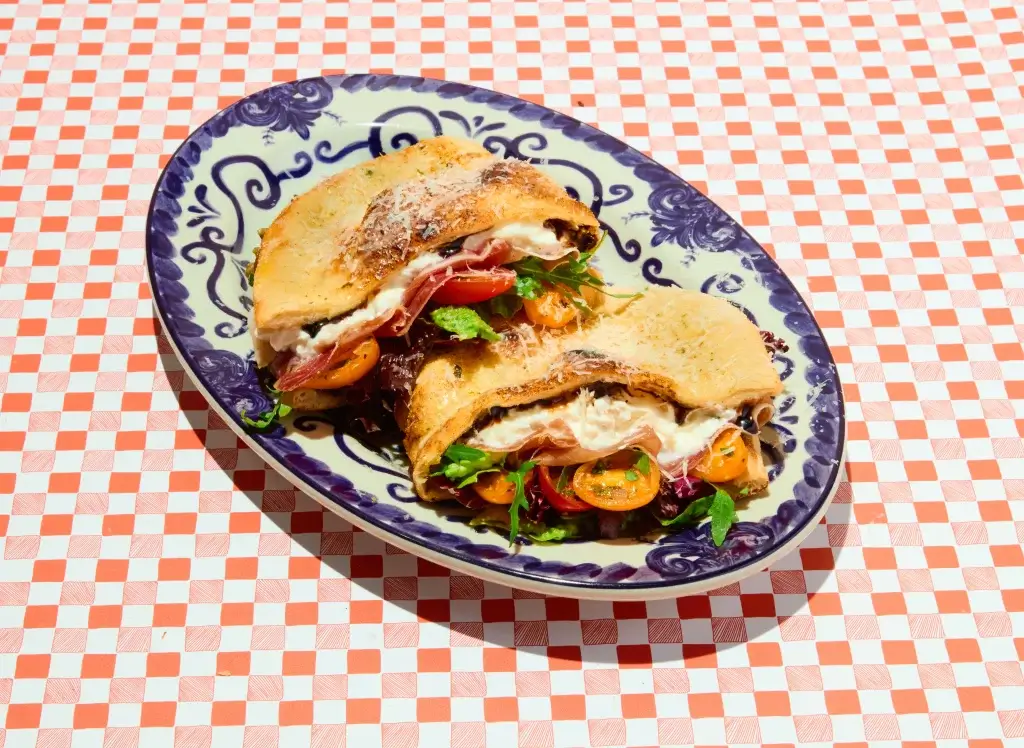 Toasted sandwich halves with melted cheese, cherry tomatoes, cured meat and salad on a blue patterned oval plate over a red-and-white check tablecloth