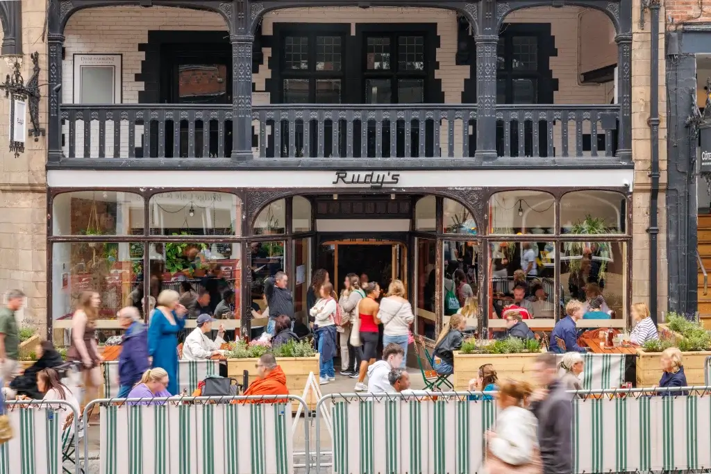 Busy outdoor seating area at Rudy's restaurant with people dining and walking in front of a black and white historic building.