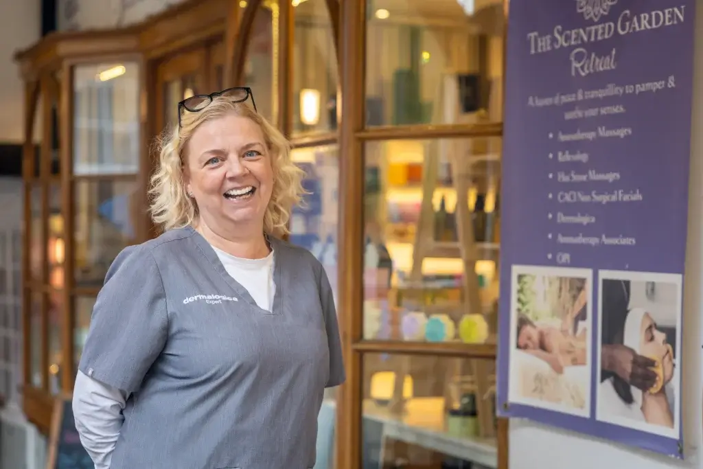 Smiling blonde woman in grey Dermalogica uniform with glasses on her head standing in front of a spa display and poster.